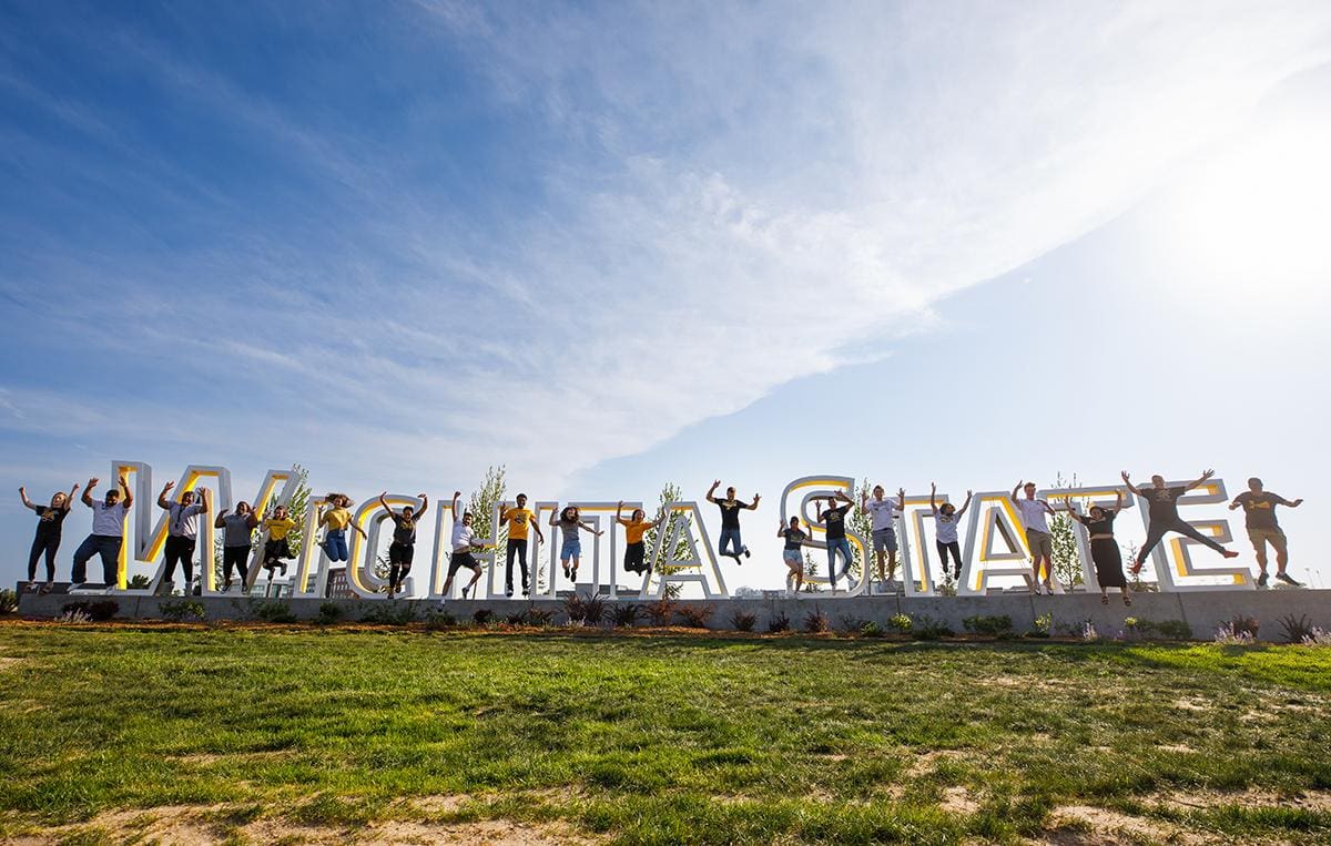 A daytime photo of a number of students standing and jumping among the illuminated letters of the Wichita State University sign at the corner of Oliver and 21st. 