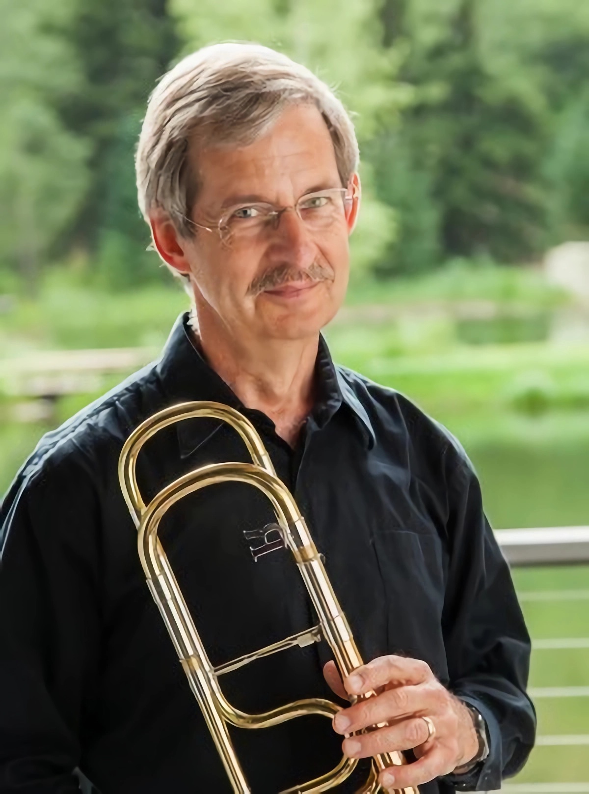 A portrait of Michael Powell. He is standing, holding his trombone with greenery in the background.
