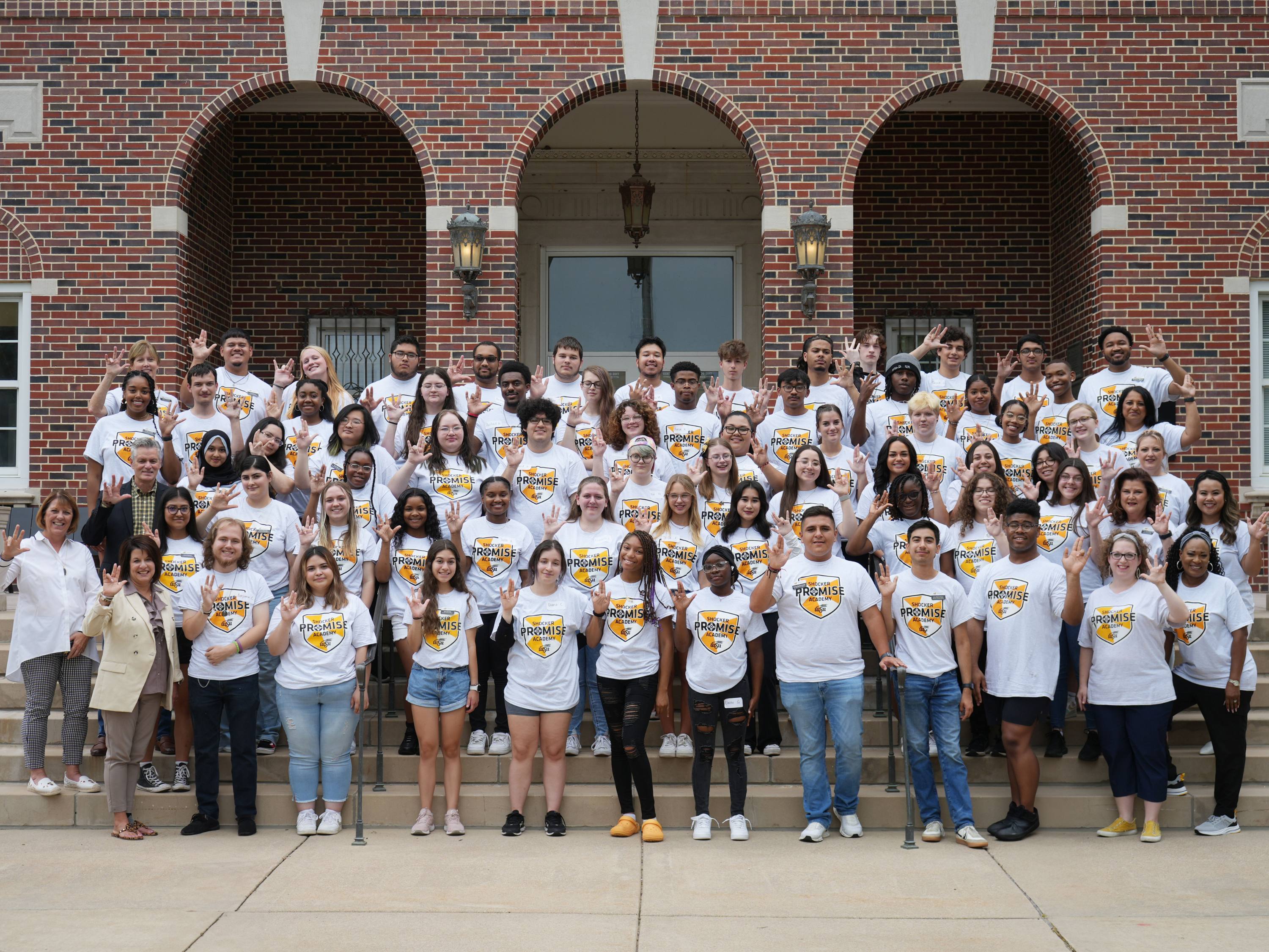A group photo of the Shocker Promise Academy Scholars posing in front of a WSU building outdoors. All of the students are wearing the same white Shocker Promise T-shirt. Among the students is WSU president Rick Muma and Financial Aid & Scholarships executive director, Sheelu Surender.