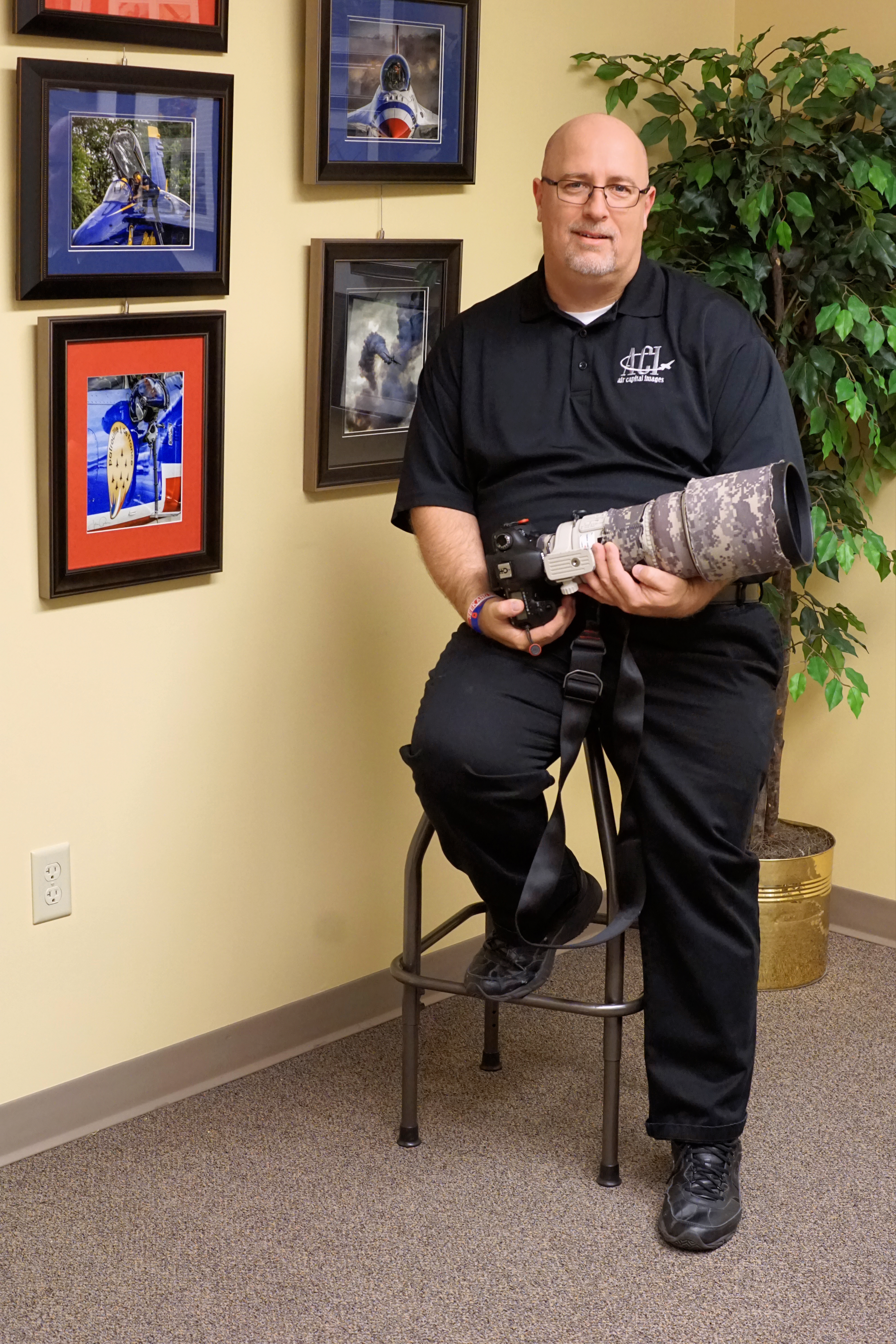 Profile photo 1 of John Gabor, sitting with his camera, surrounded by displayed photos he has taken. 