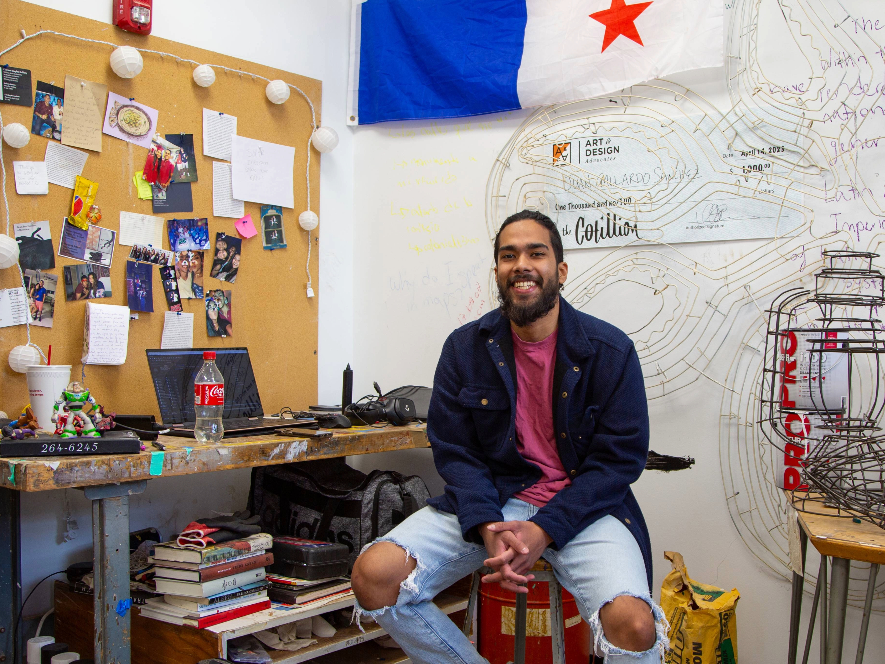 MFA student Juan Gallardo Sánchez sits in a chair in his Henrion Hall studio surrounded by art supplies, wire sculptures, and electronic equipment.