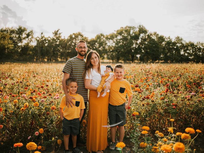 Merry Kirkpatrick stands in a field of yellow flowers with her 3 kids. One is a little baby girl in her arms. The other two are little boys. Her husband stands behind her. They are all wearing yellow themed clothes to match the flowers. There are some trees that line the blue sky.