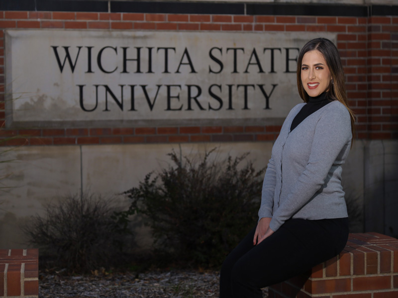 Valeria Esparza sits in front of the Wichita State University sign. She is wearing a grey sweater and black turtleneck and is sitting on a brick ledge. There is some shrubbery below the sign. 