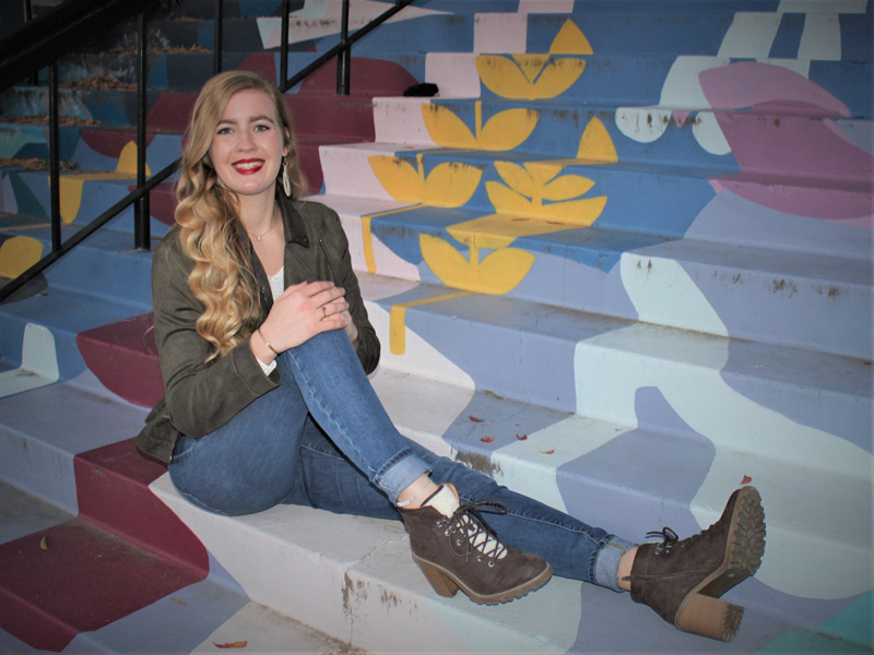 Lysel Bender is wearing a dark green jacket, some jeans, and brown high heel boots. She is sitting on the steps of the Barton School of Business where the purple, blue, yellow, and pink mural is.