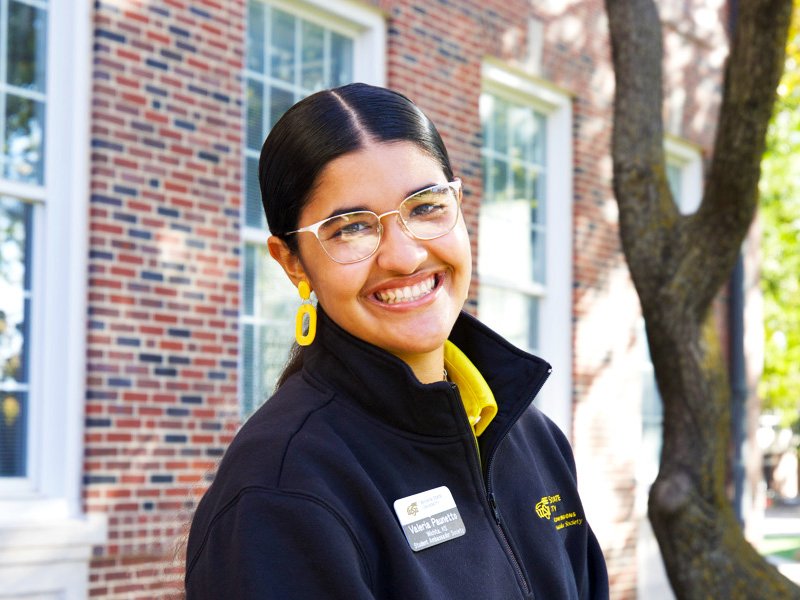 Valeria Paunetto is standing in front of the brick laid Morrison Hall and there is a tree in the background. The sun is shining. Paunetto is wearing her shocker gear with some yellow earrings and she is smiling. 