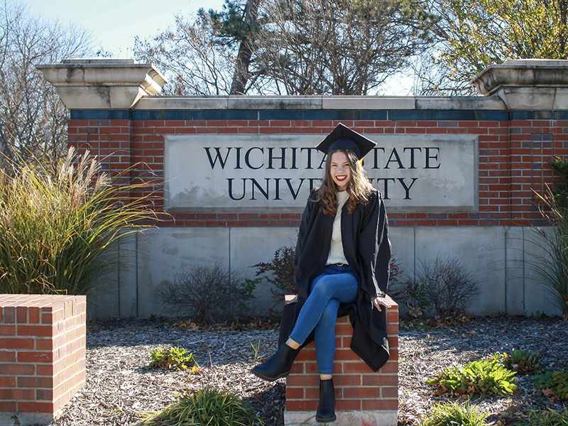 Kennedy Stamm sits in from of WSU sign at the entrance with the two columns by it. She is in her grad cap and stole. There is some nice shrubbery in the background. She is smiling and sitting on a brick chair/ 