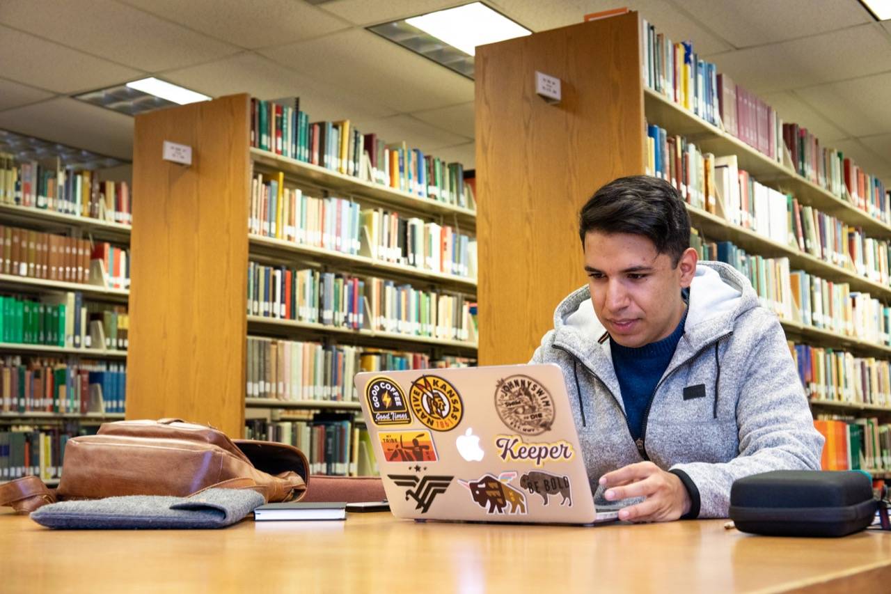 Photo of a student working on his laptop with library shelves in the background. 