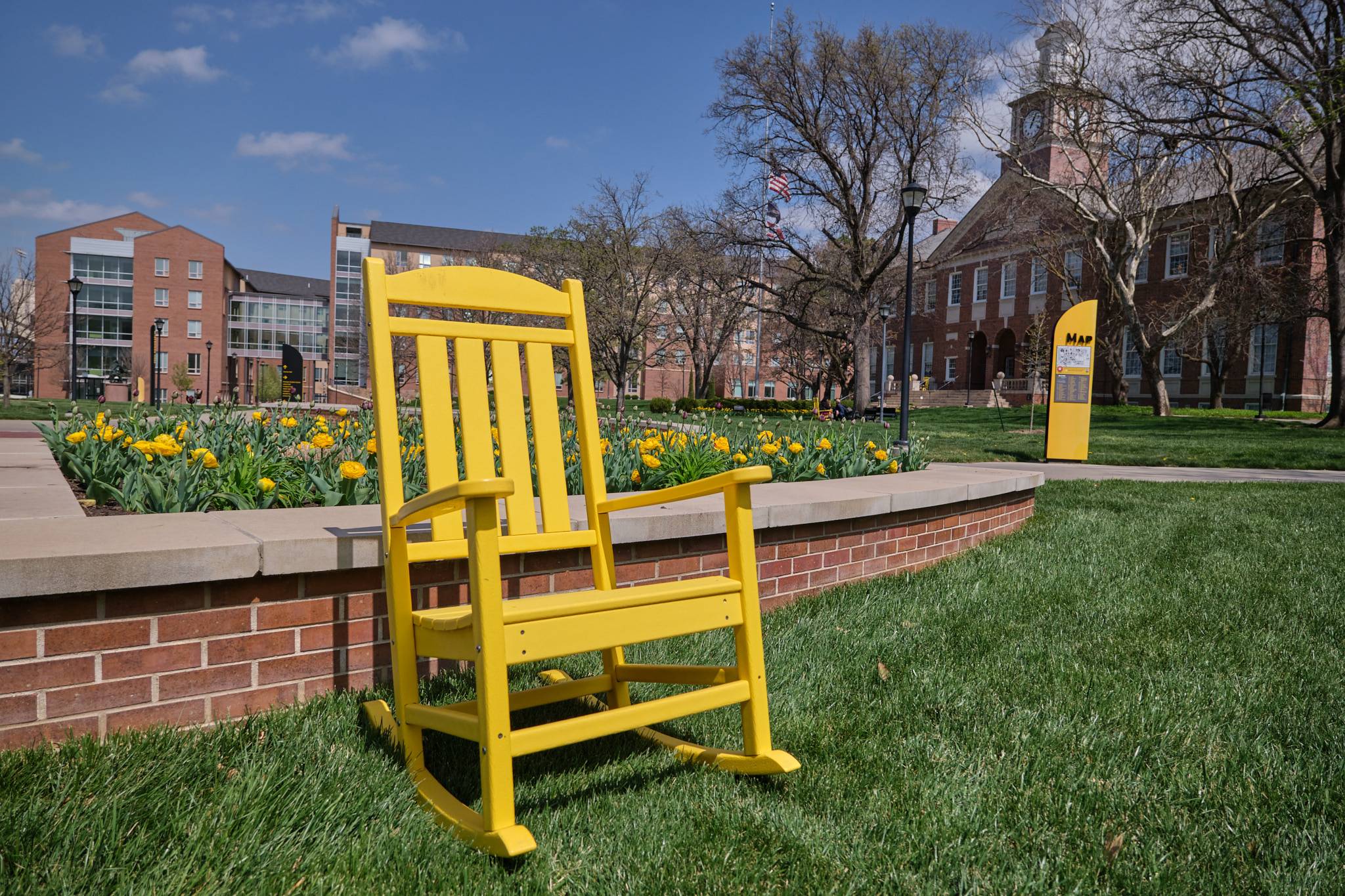 an empty rocking chair sits on green grass in front of a flower bed 