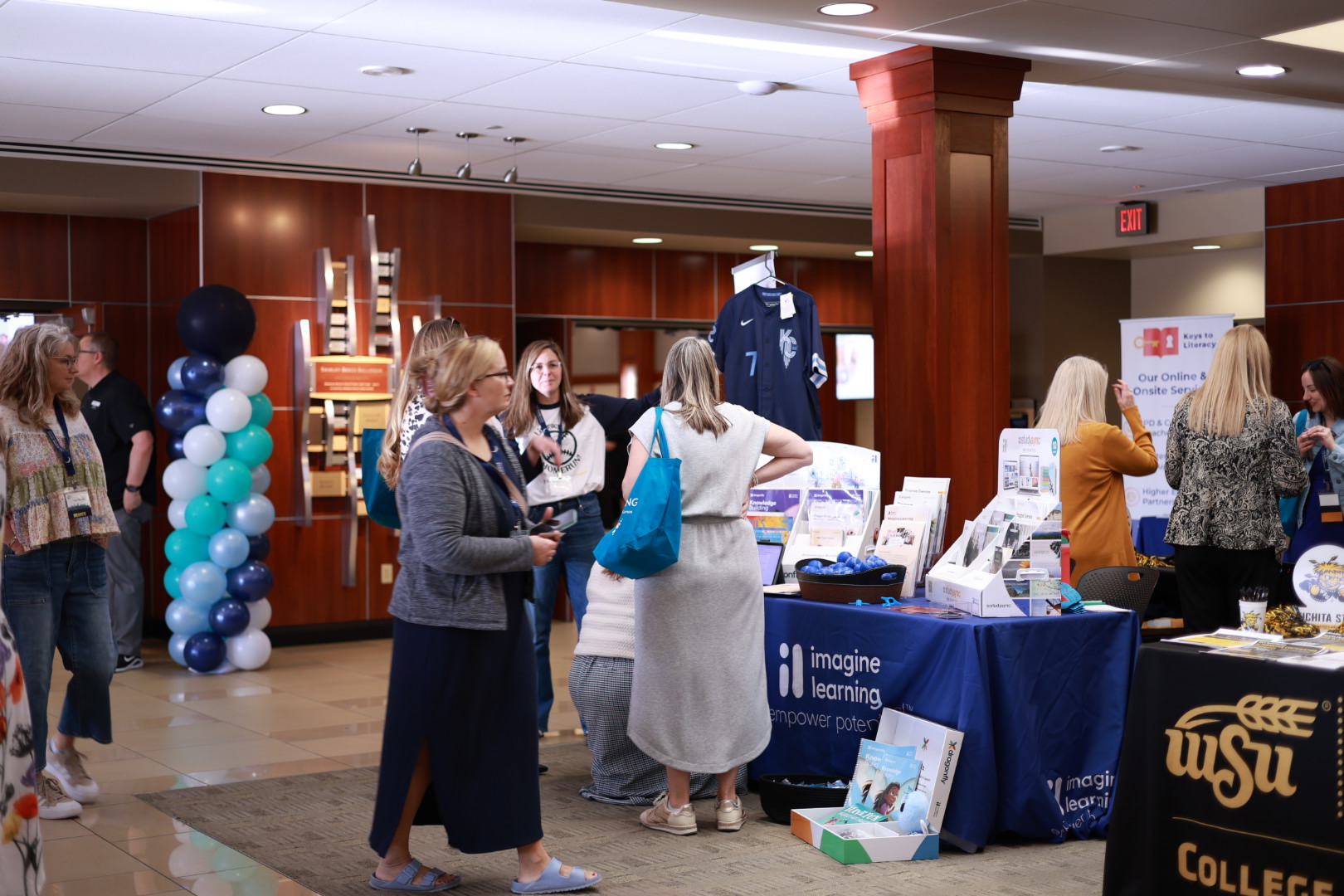 Participants walk through a conference area with vendor and information tables set up. Displays, books and Wichita State materials are visible as attendees browse and connect.