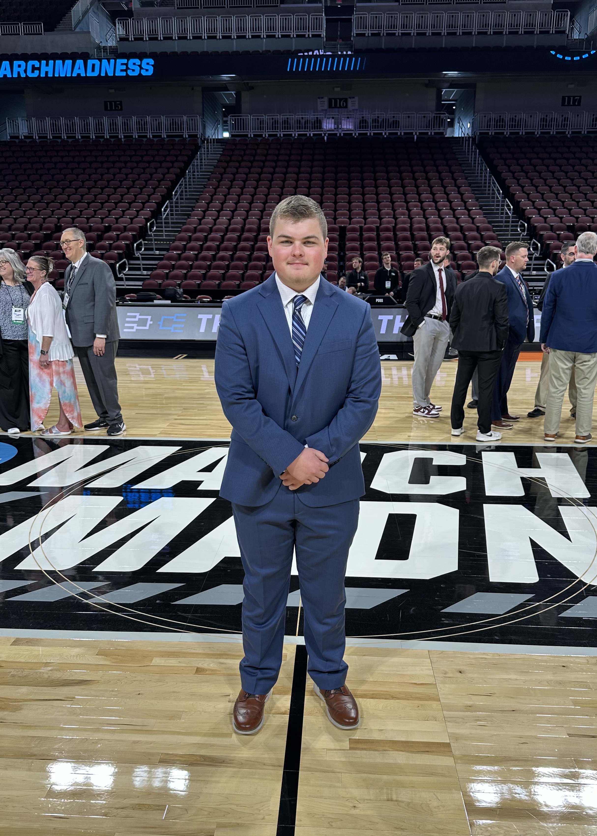 WSU student Liam Crawford stands on the court at the NCAA tournament venue in a navy suit and striped tie. The March Madness logo is visible beneath his feet, with event staff and attendees in the background.