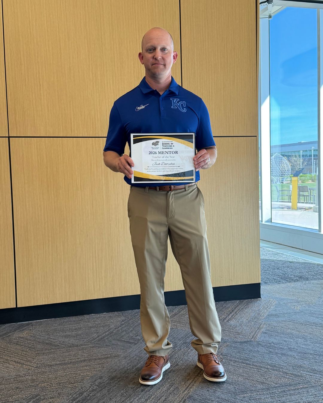 A man stands indoors holding a “2026 Mentor Teacher of the Year” certificate from Wichita State University’s School of Teaching and Learning. He is wearing a blue polo shirt with a KC logo, khaki pants and brown shoes, with a window and campus view to the side.