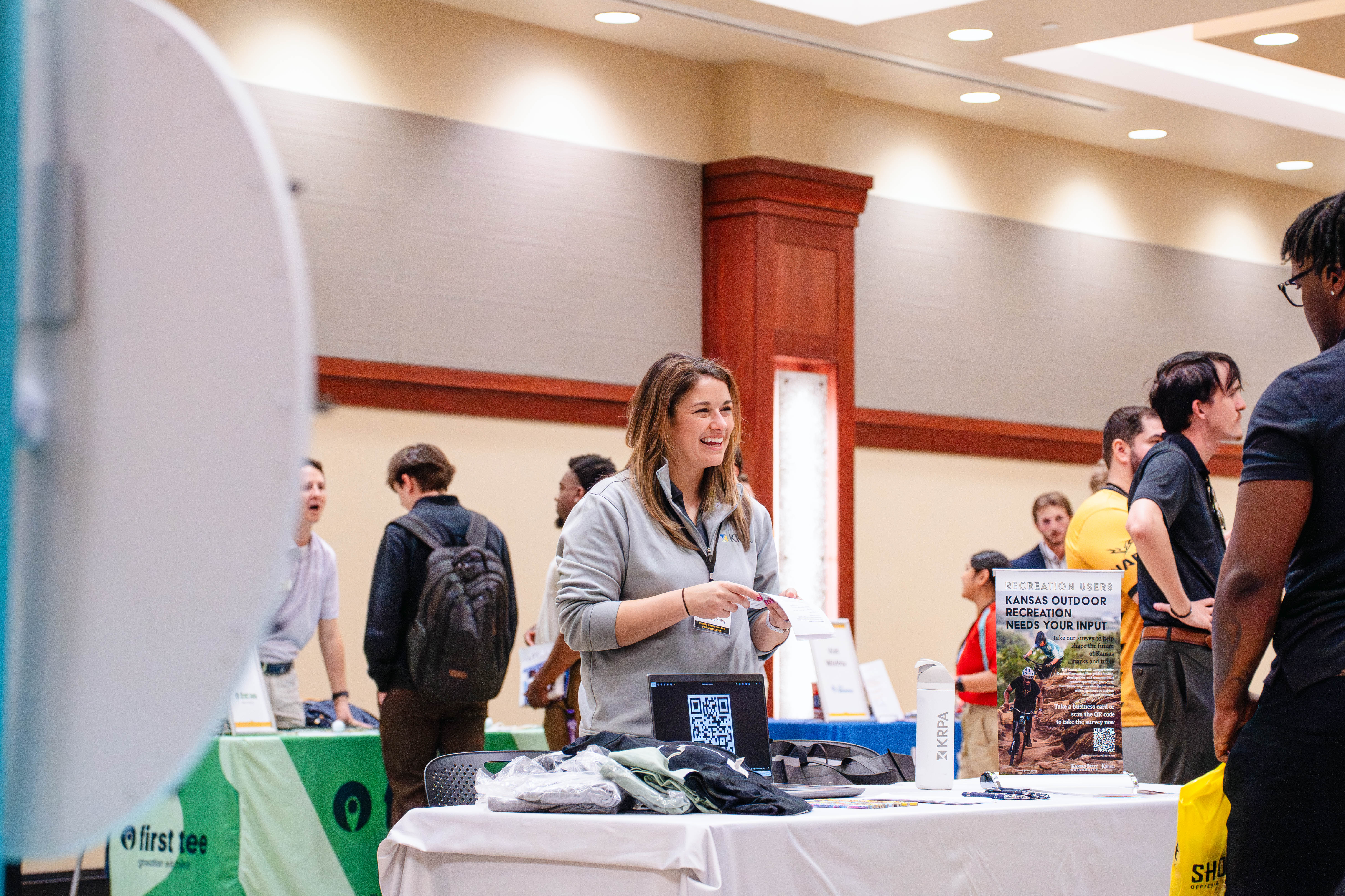 A woman speaks with attendees at a booth during the Kansas Sport Summit at Wichita State University, standing behind a table with informational materials and a QR code display.