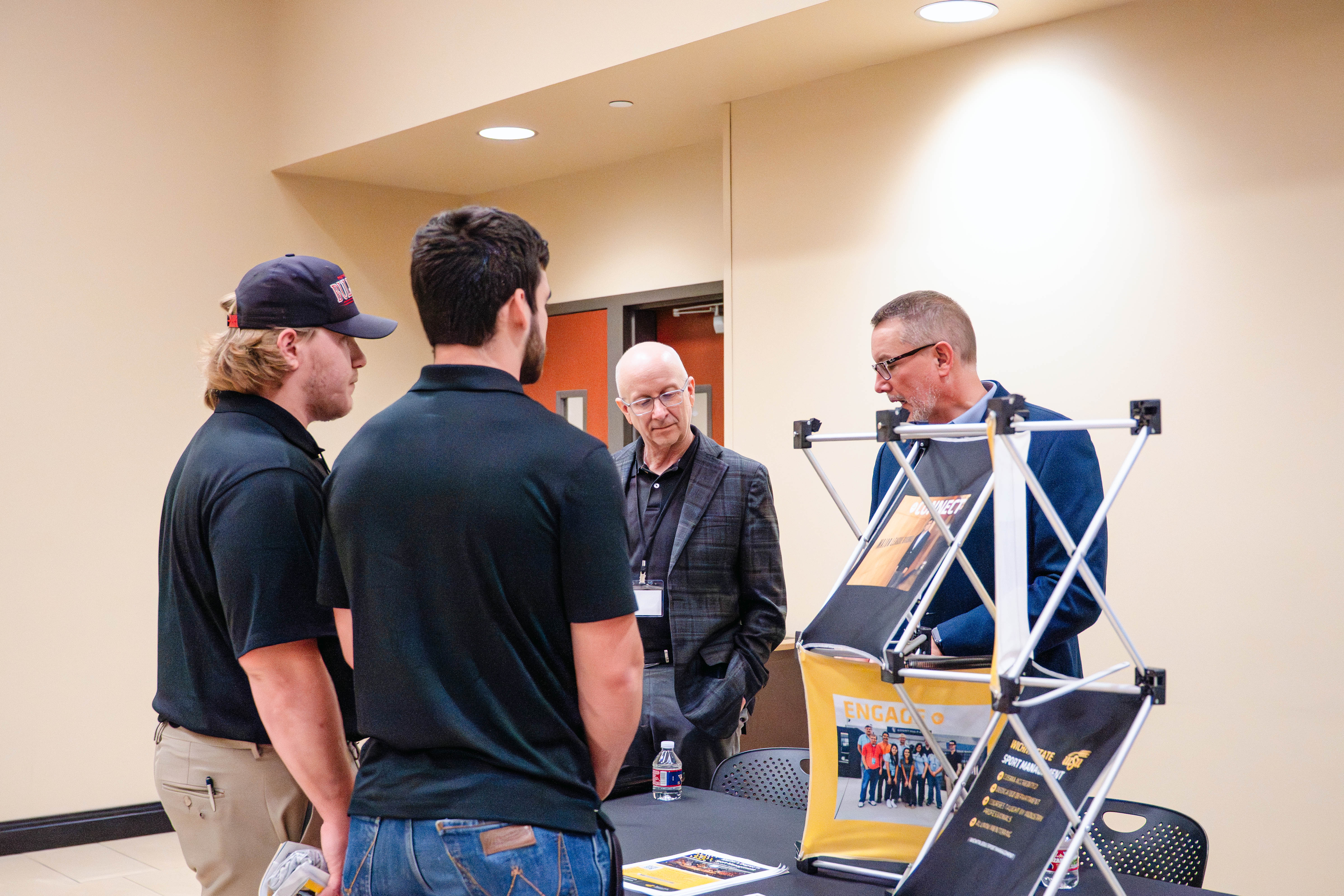 Students speak with Wichita State sport management staff at a booth during the Kansas Sport Summit at Wichita State University, gathered around a table with informational materials in a conference room.
