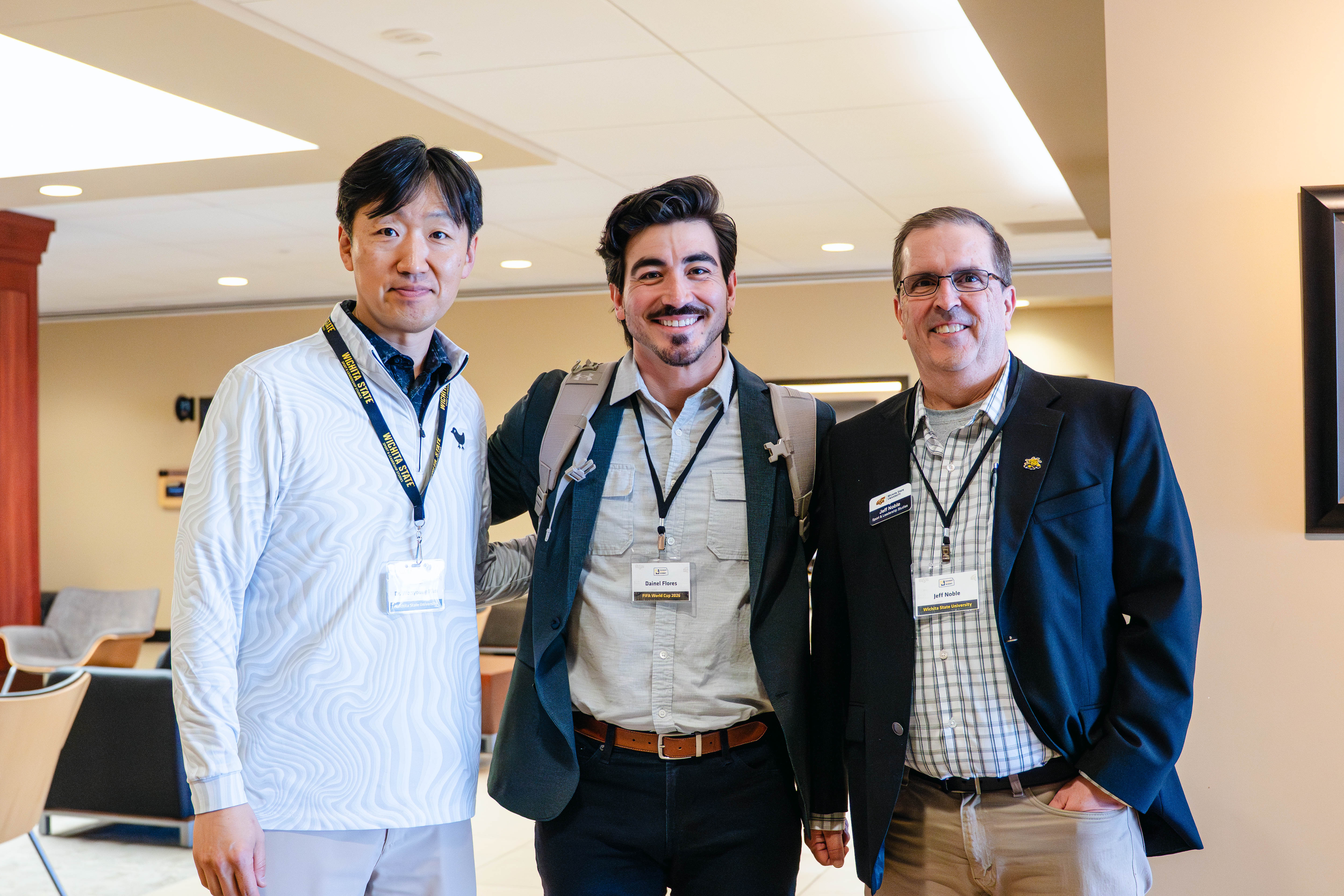 Two men review information at a booth during the Kansas Sport Summit at Wichita State University, looking down at materials together in a conference room setting. Three men pose together at the Kansas Sport Summit at Wichita State University, smiling and wearing name badges in a lobby area.