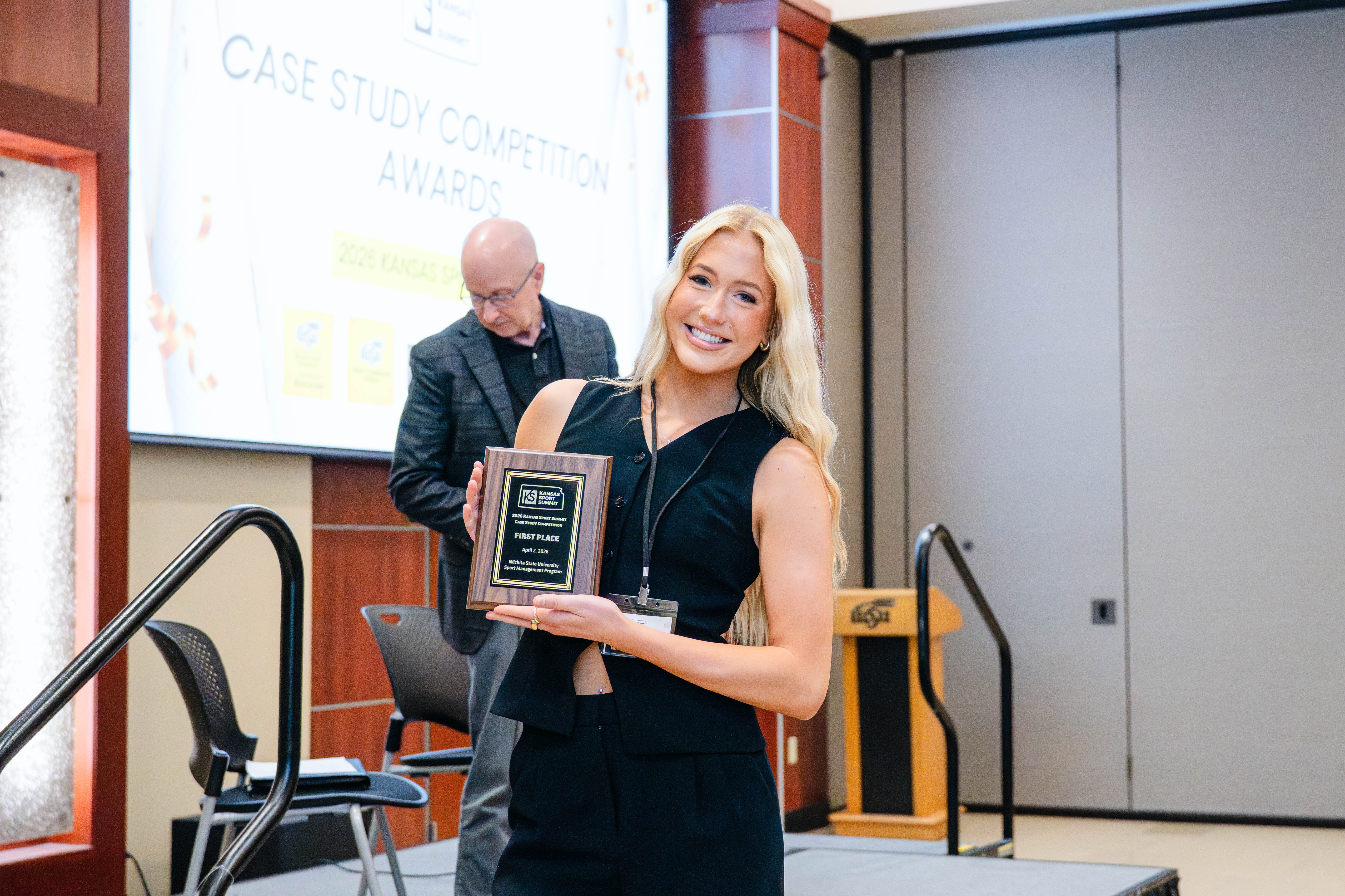 A student smiles while holding a first-place award plaque during the case study competition at the Kansas Sport Summit at Wichita State University.