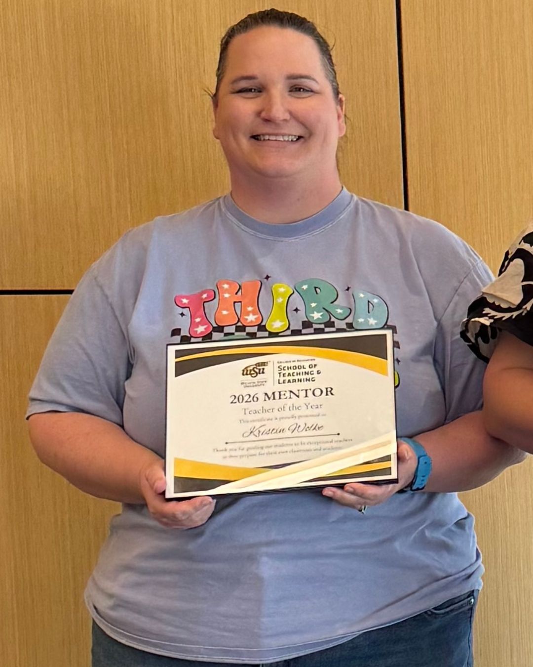 A woman stands indoors holding a “2026 Mentor Teacher of the Year” certificate from Wichita State University’s School of Teaching and Learning. She is wearing a gray T-shirt and smiling at the camera.
