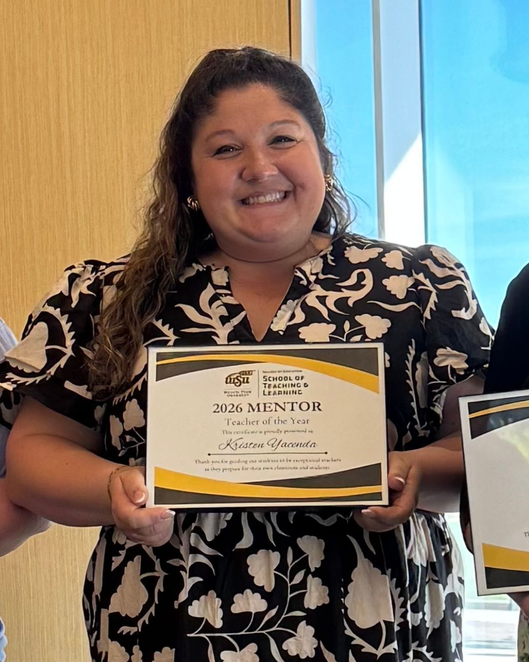 A woman smiles while holding a “2026 Mentor Teacher of the Year” certificate from Wichita State University’s School of Teaching and Learning. She is wearing a patterned dress and standing indoors with part of another person visible beside her.
