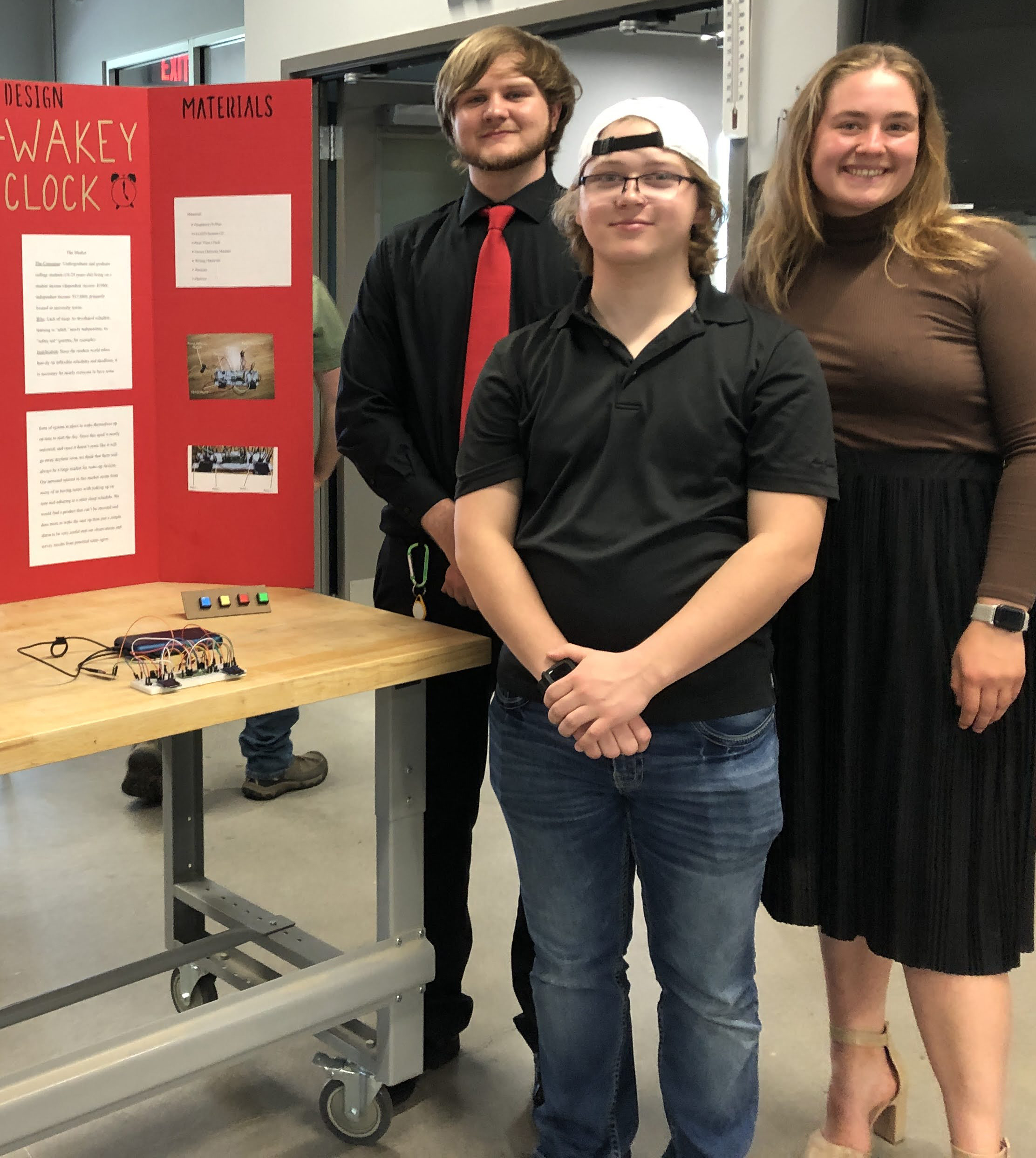 A team of students proudly stands next to their project display board and prototype.