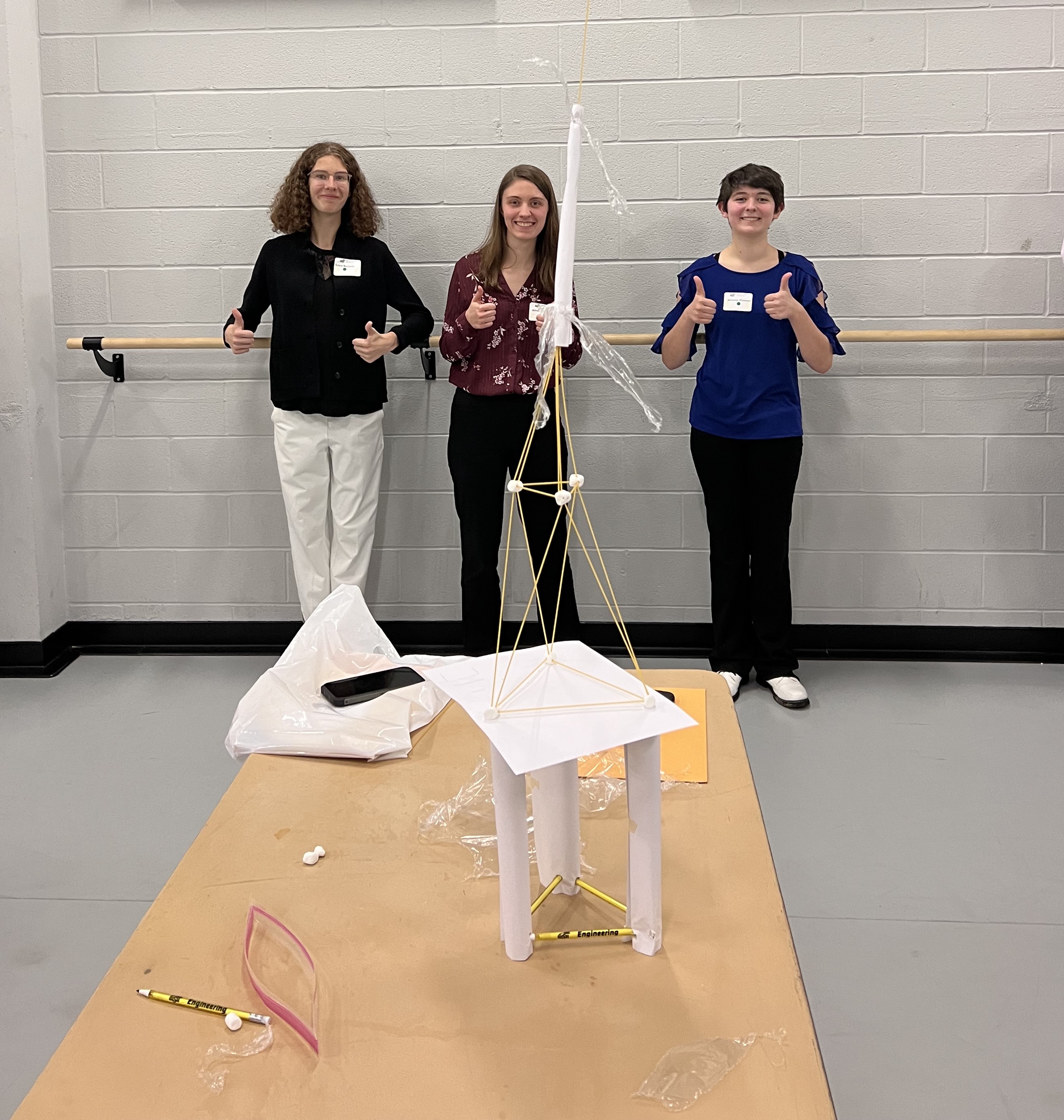 A group of high school women stands behind their large spaghetti marshmallow tower giving a big thumbs up.