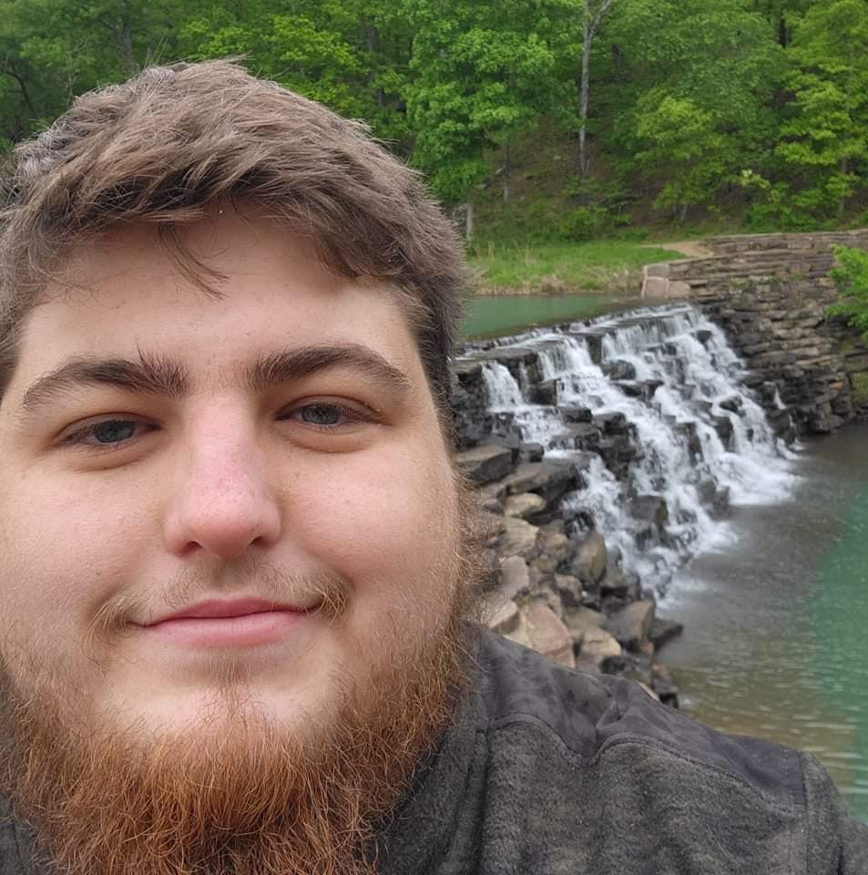 smiling face of a young white man with beard in front of a waterfall