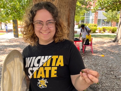 Student smiling at the camera while holding a bug swooping net in one hand and a beetle in the other.