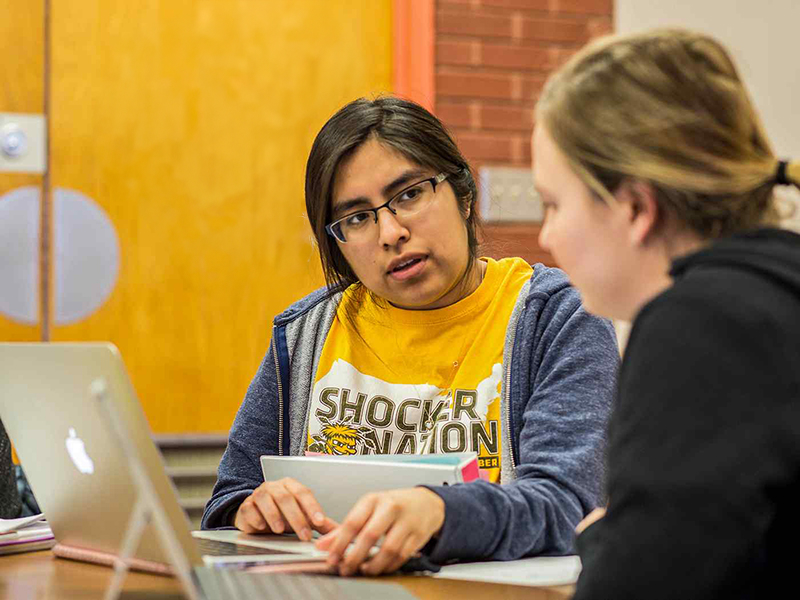 Two students discuss an assignment in front of their laptops