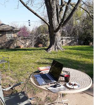 Photograph showing a laptop computer on an outdoor patio table with an empty desk chair at the table as if someone has been working and is now absent. The scene is set outdoors during a sunny day. 