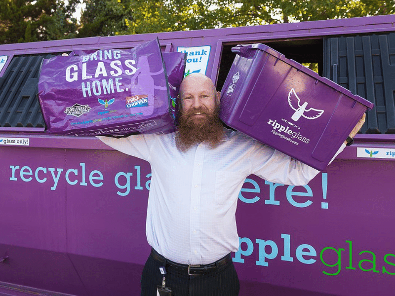 Man standing holding two bins of recyled glass on his shoulders standing in front of a Ripple Recycling bin.
