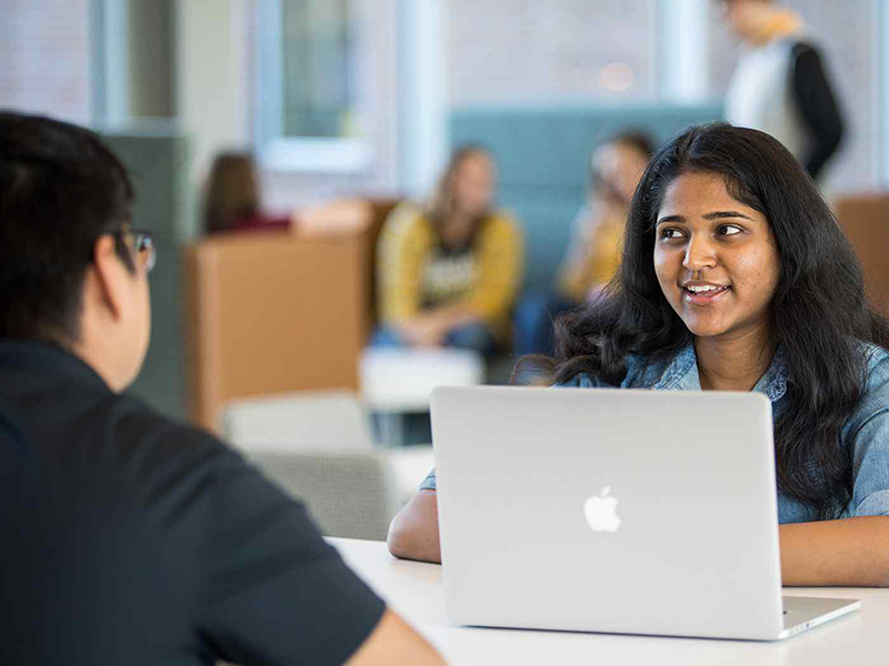 A student with a laptop talks to another student