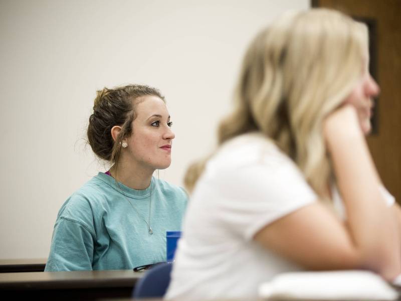 Female student listens attentively in a classroom, with another student blurred in the foreground.