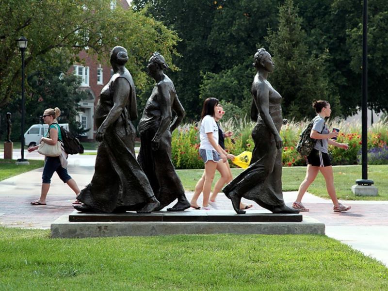 Bronze statue of three women walking, titled 'Tres Mujeres Caminando' ('Three Women Walking'), depicting prehistoric-inspired women, displayed alongside students walking on a path at Wichita State University.