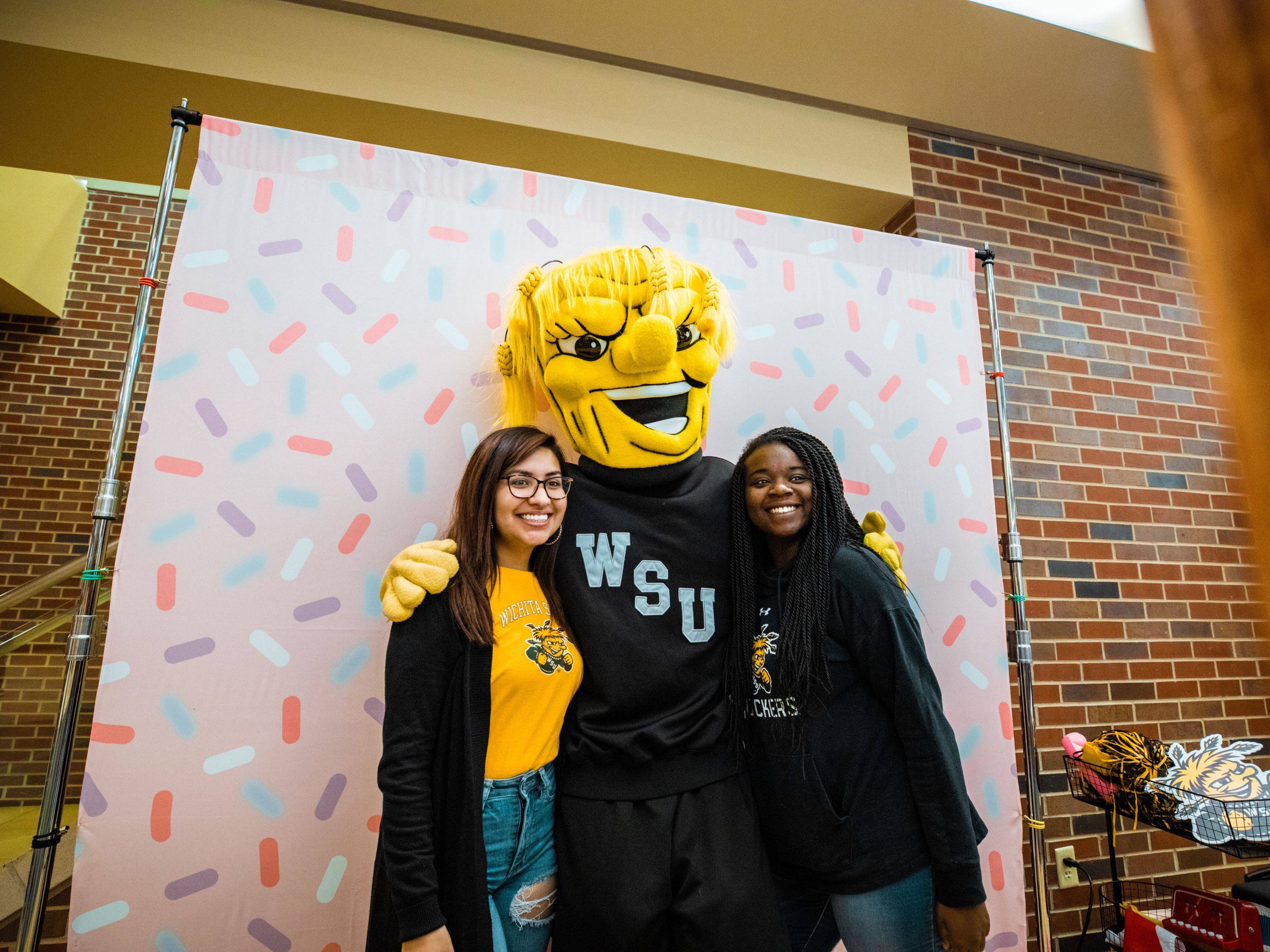 Two female event participants pose for a photo with Wichita State's mascot, Wu, in front of a colorful backdrop.