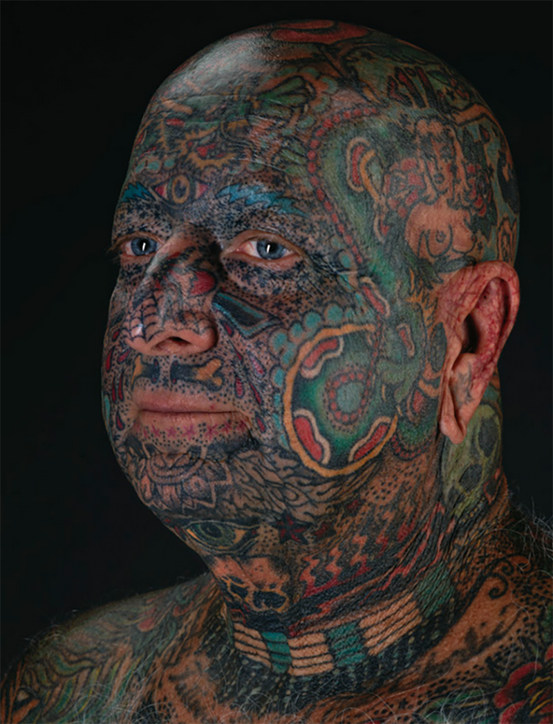 Portrait photograph of a bald man, his face covered in tattoos.