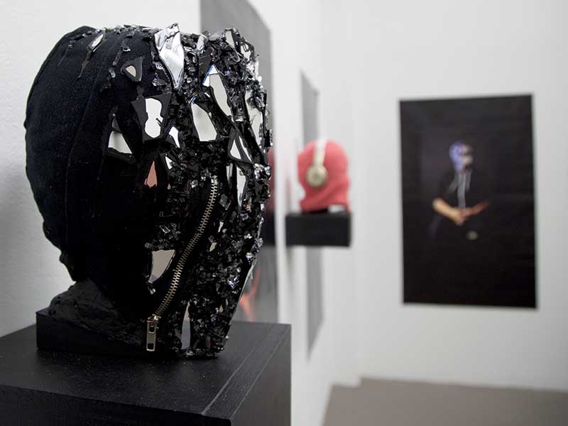Photograph of works by Alexis Rivierre in the ProjectSpace gallery. A black mask, covered with small pieces of jagged, mirrored glass dominates the foreground. A pink crochet mask with silver earphones and a photograph of the artist wearing a mask made out of an American flag are pictured in the background. 