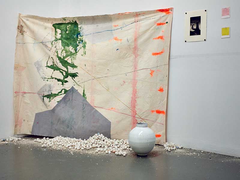 Photograph of a student installation in the ProjectSpace gallery: a large abstract painting on a piece of unstretched canvas hangs across the cover of a wall behind a white ceramic vase, and white ceramic objects that look like rocks. A black-and-white photograph and two small paintings, one pink and one yellow, hang by the canvas.