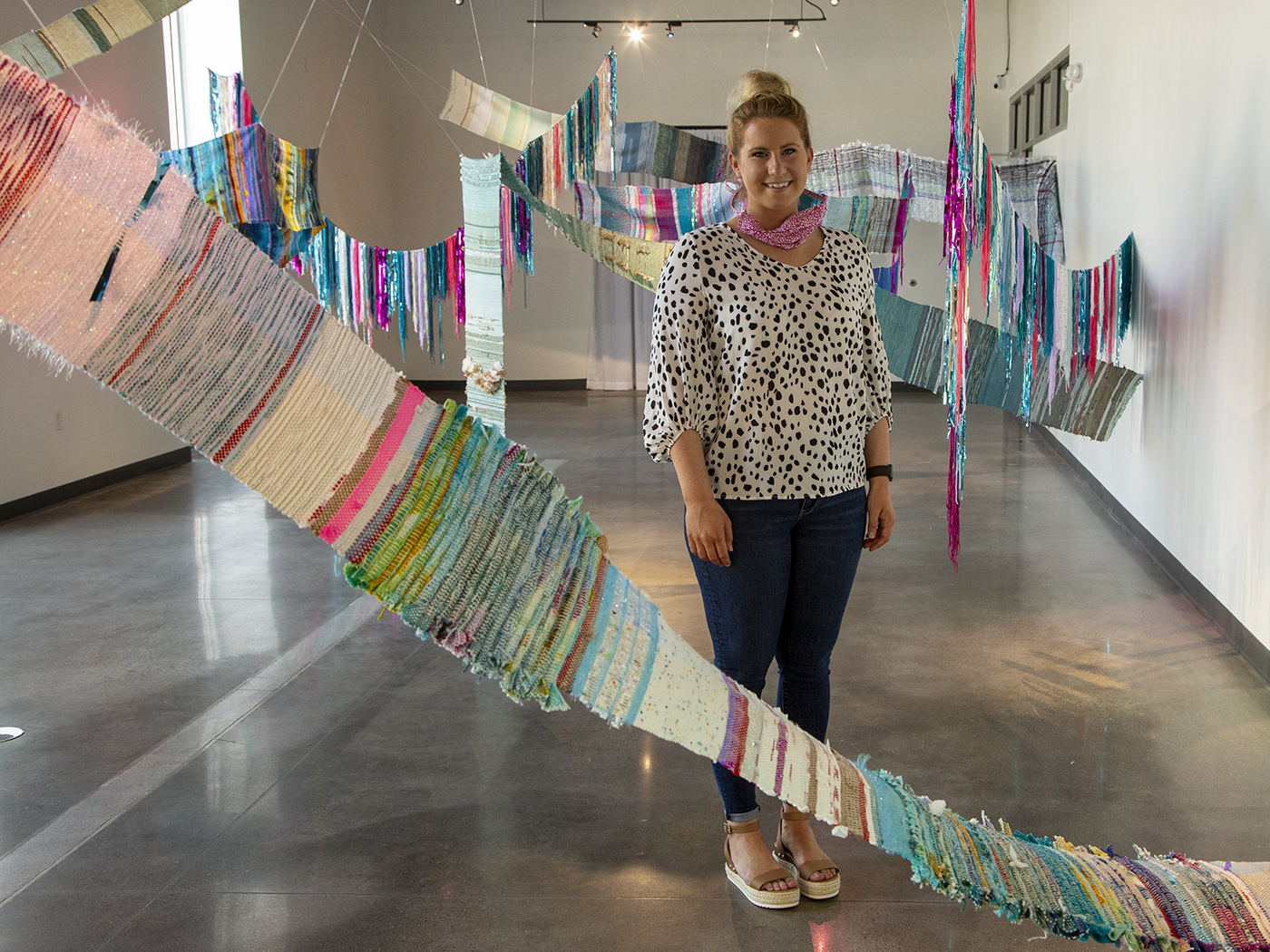 Amy Huser stands in the middle of "Casually Not Okay," her Master of Fine Arts thesis exhibition. She is surrounded by brightly colored weavings that are hung from the ceiling.