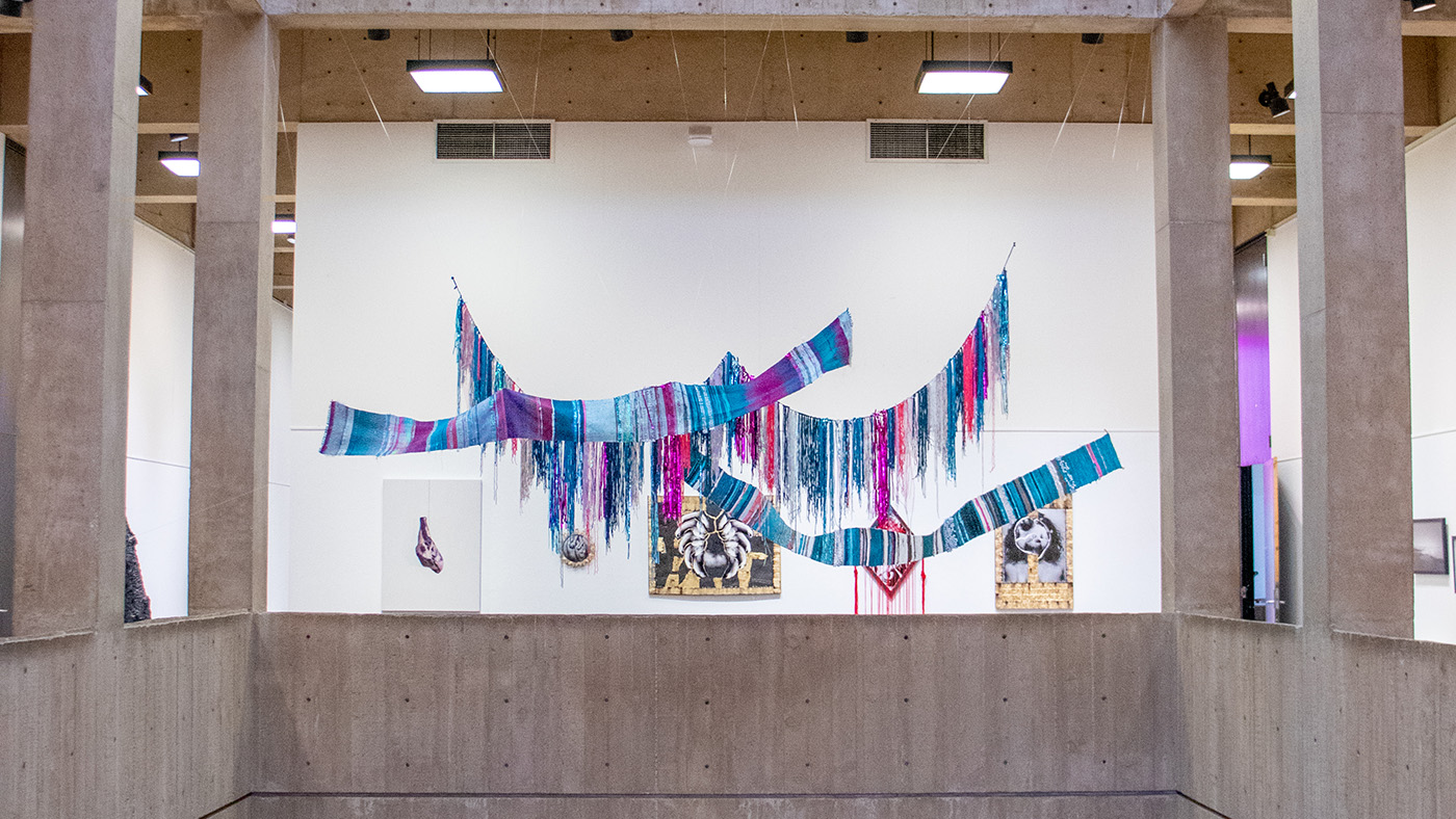 A large woven installation hangs in the Atrium of McKnight Art Center, in front of five two-dimensional works.