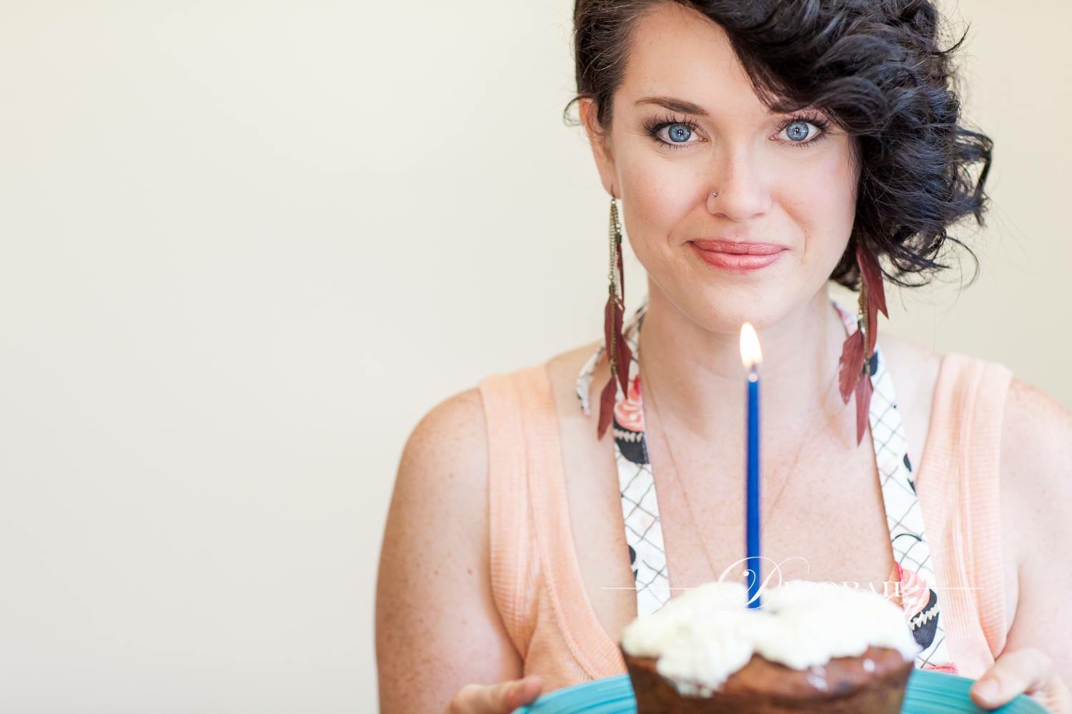 Promotional photo of Cassandra Nuss, smiling, offering a cupcake with a candle to the viewer. 