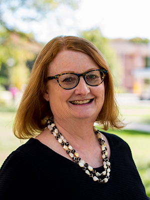 Dr. Elaine Bernstorf posing for a headshot photo in a black shirt, black glasses, and a black/white necklace. The background is blurred.