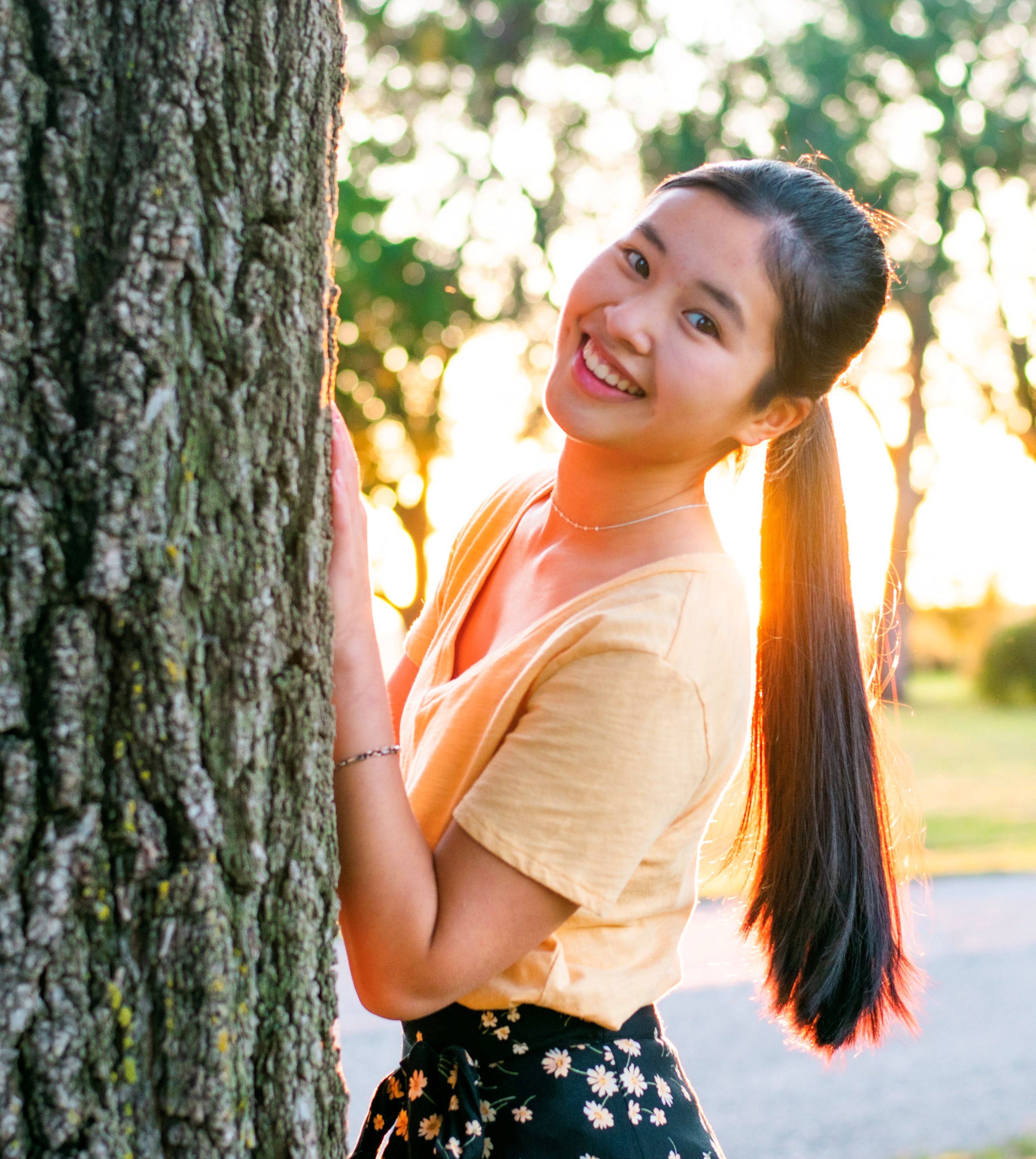 Honors student Jaden Wood posing for a headshot photo. Jaden has one hand placed on a tree, the sun setting behind her. She is wearing a yellow shirt with black bottoms and her hair is in a ponytail.