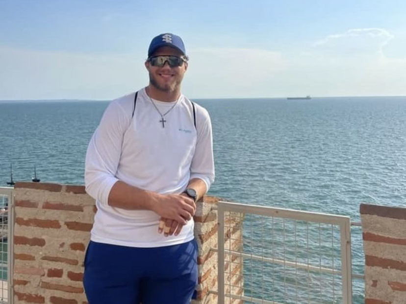 Honors student Alex Sterzing posing for a photo in Greece. Alex is wearing a white long-sleeve shirt, blue shorts, a blue hat, and black sunglasses. He is leaning against a brick fence with the ocean and a ship behind him.