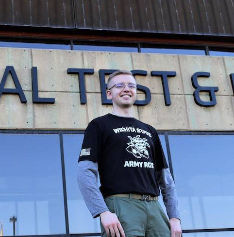 Honors student Cameron Holston posing for a photo outside NIAR's Aircraft Structural Test and Evaluation Center. Cameron wears a black Wichita State Army ROTC t-shirt and green pants. He is blonde and wears wire-rimmed glasses.