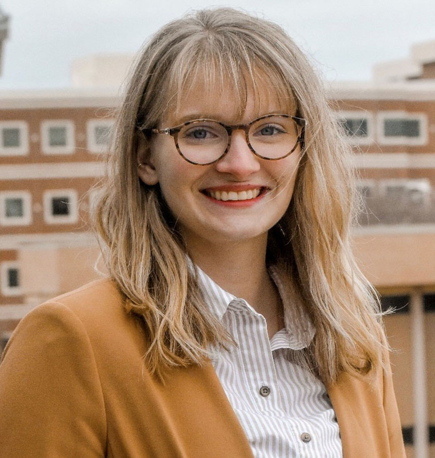 Honors student Hanna Chastain posing for a headshot. Hanna is wearing a dark beige suit jacket over a white and light brown striped button-up shirt and multi-colored glasses. She is blonde with blue eyes and is smiling while looking directly at the camera. The background of the photo is blurred.