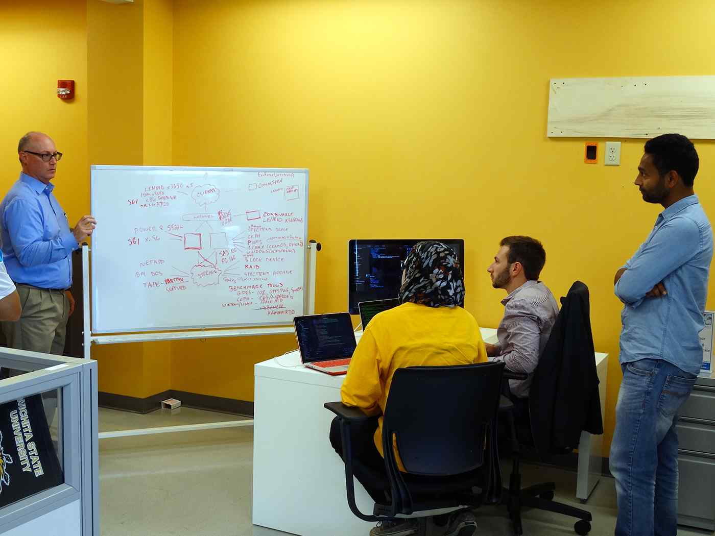 Computer Science instructor presenting lesson information on a whiteboard to several students. 