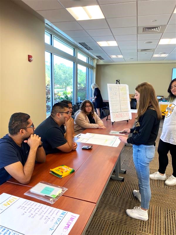 A photo of a Shocker Financial Wellness peer counselor standing in front of a group of students who are sitting down. The peer counselor is interacting with the students for a workshop activity.