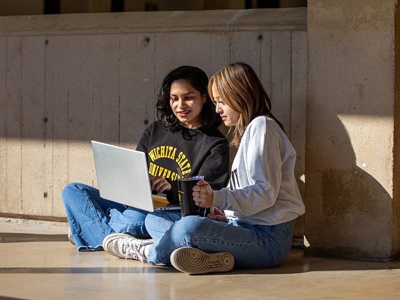 Two students sit cross-legged on the floor of McKnight Hall while they both look at a laptop