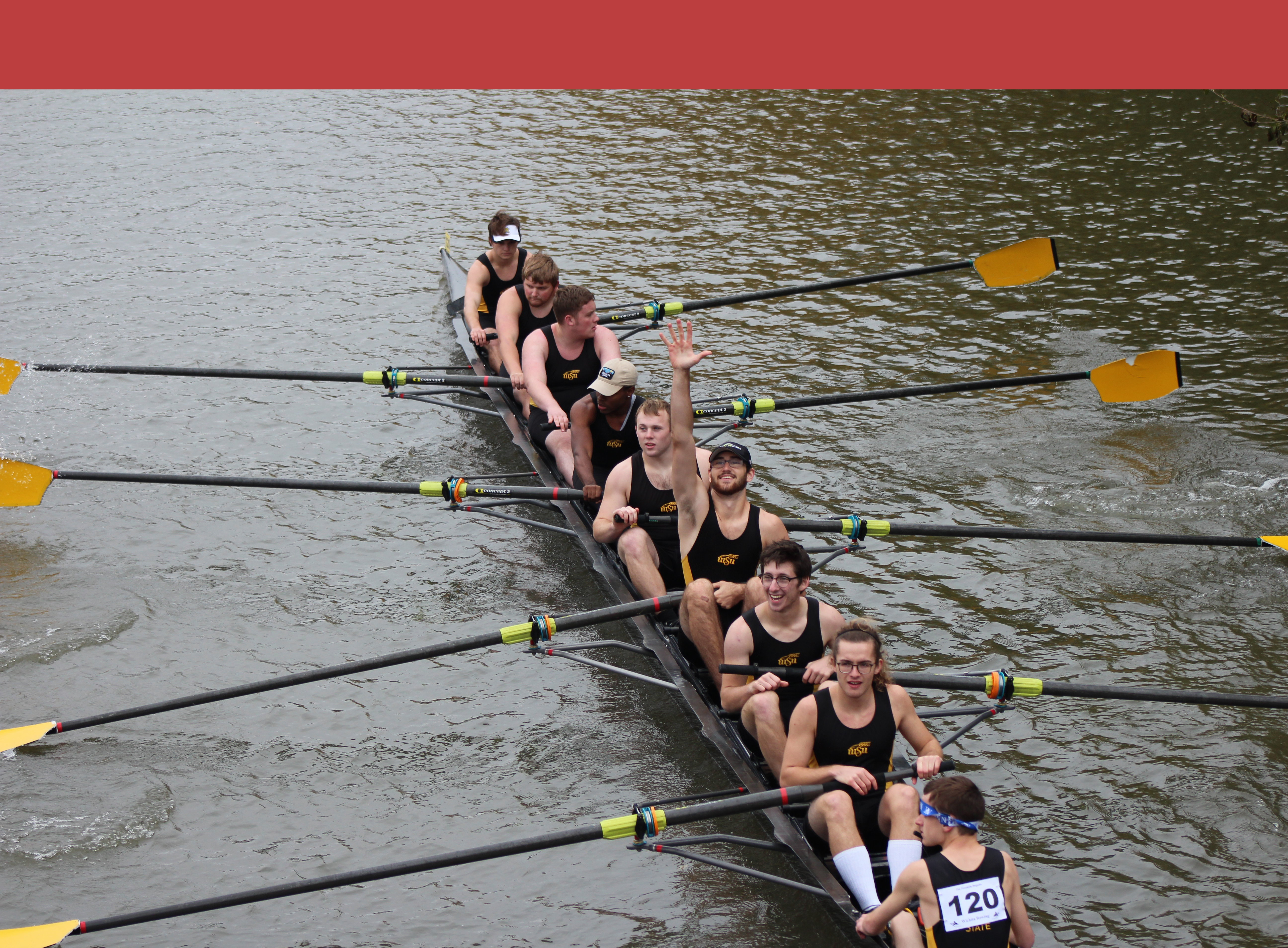 Men's novice 8. One of the men in the middle of the boat has his hand upraised and is smiling at the camera. All the rest are focused on rowing like good rowers.