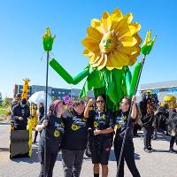 The giant Sunflower with a face and moving hands is being propped up by students who are smiling and having a good time.  