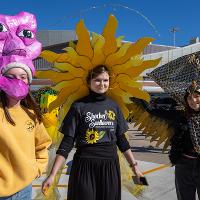 Three people show off their parade costumes. The person on the left has a purple artistic face on their head. The middle person looks like a sun or a sunflower. The person on the right has a black and yellow peacock costume.