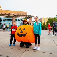 Person in an inflatable jack-o-lantern stands with two kids who are about to run the Pumpkin Run race. 